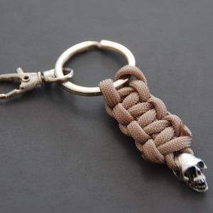 Peut inclure: Un porte-clés argenté avec un cordon en paracorde brun enroulé autour de l'anneau et un charme en forme de crâne argenté attaché à l'extrémité.
