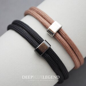 Könnte beinhalten: Zwei Armbänder mit verstellbaren Metallverschlüssen. Ein Armband ist schwarz und das andere ist hellbraun. Die Armbänder bestehen aus einem geflochtenen Kordelmaterial.