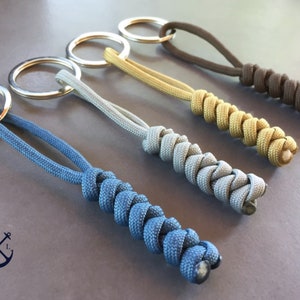 Peut inclure: Cinq porte-clés avec des cordons tressés de différentes couleurs attachés à des anneaux de clés argentés. Les cordons sont bleus, gris, dorés et bruns.
