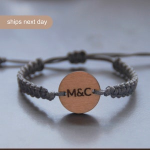以下が含まれることがあります： 「M&C」と刻まれた丸い木製のチャームが付いたグレーの編み込みブレスレットです。