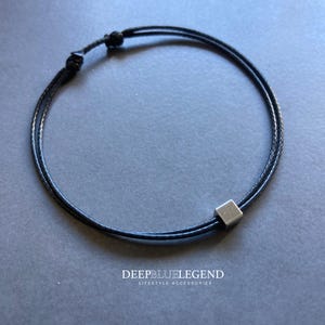 Puede incluir: Pulsera de cordón negro con una pequeña cuenta cuadrada de color plateado. La pulsera es ajustable con un cierre de nudo corredizo. El texto "DEEPBLUELEGEND LIFESTYLE ACCESSORIES" está en la parte inferior de la imagen.