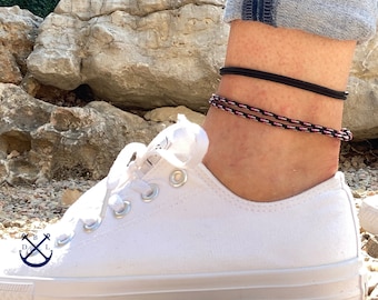 Può includere: Braccialetti alla caviglia. L'immagine mostra due braccialetti su una caviglia. Uno è nero, l'altro è una combinazione di nero, rosa e bianco. Una scarpa di tela bianca è in primo piano. Lo sfondo è un ambiente naturale all'aperto.