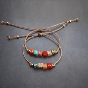May include: Two adjustable bracelets with colorful beads spelling out the names "Doree" and "Mama". The bracelets are made with brown cord and gold beads.