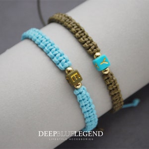 May include: Two friendship bracelets with personalized letter charms. The bracelet on the left is light blue with a gold "E" charm. The bracelet on the right is olive green with a turquoise "Y" charm.  DeepBlueLegend Lifestyle Accessories.
