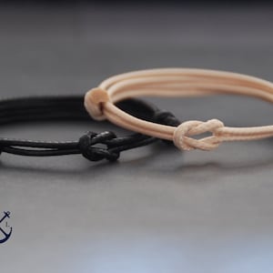 May include: Two minimalist bracelets. One is black, the other is beige. Both are made of cord and feature a knot closure. The bracelets are displayed on a dark gray surface. The bracelets are simple and elegant.
