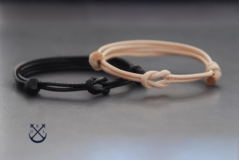 May include: Two minimalist bracelets. One is black, the other is beige. Both are made of cord and feature a knot closure. The bracelets are displayed on a dark gray surface. The bracelets are simple and elegant.
