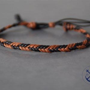 Puede incluir: Un brazalete trenzado con cordón negro y marrón. El brazalete está atado con un nudo en cada extremo.