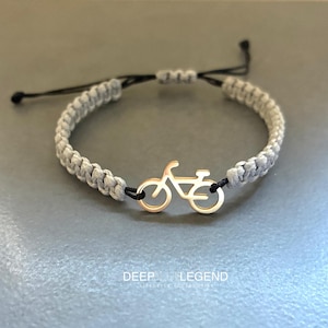 Op de afbeelding: Een grijze gevlochten armband met een goudkleurige fietsbedel. De armband heeft zwarte verstelbare koorden en een fietsvormige hanger. De tekst "DEEP LEGEND" staat onderaan de afbeelding.