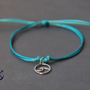 May include: A turquoise blue adjustable string bracelet with a silver charm featuring a wave design.