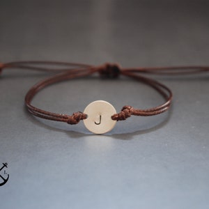 Peut inclure: Un bracelet en cordon marron avec un petit charme rond en métal gravé de la lettre "J".