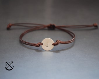 Puede incluir: Un brazalete de cordón marrón con un pequeño dije redondo de metal con la letra "J" grabada.