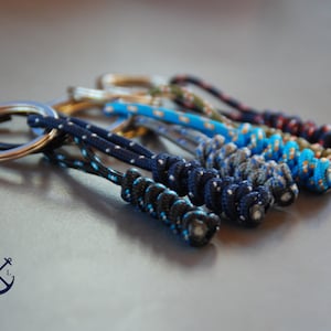 Op de afbeelding: Een set van zes kleurrijke sleutelhangers gemaakt van gevlochten touw met een zilveren sleutelring. De sleutelhangers zijn in tinten blauw, groen en rood.
