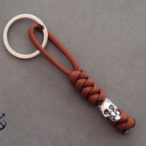 May include: A brown paracord keychain with a silver skull bead. The keychain is attached to a silver key ring.