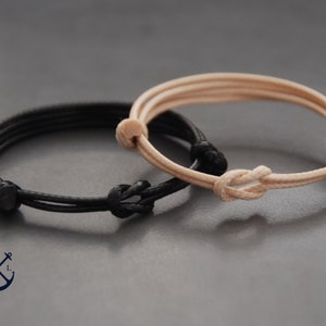 May include: Two adjustable bracelets. One is black, the other is tan. Each bracelet features a knotted design and is made of a thin, cord-like material. The bracelets are displayed on a gray surface.