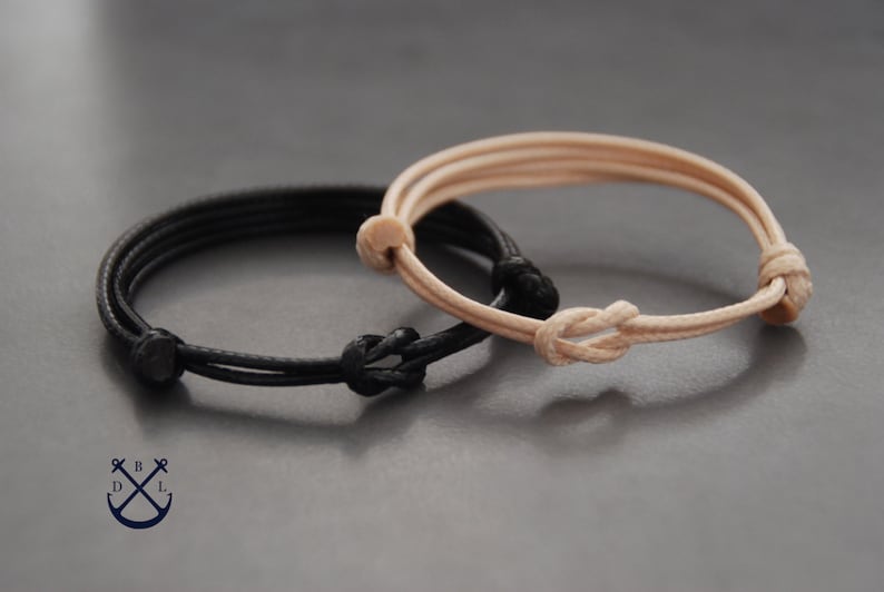 May include: Two adjustable bracelets. One is black, the other is tan. Each bracelet features a knotted design and is made of a thin, cord-like material. The bracelets are displayed on a gray surface.