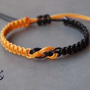 Könnte beinhalten: Ein schwarzes und goldenes Freundschaftsarmband mit einem Unendlichkeitsknotendesign. Das Armband ist aus geflochtener Schnur gefertigt und hat eine verstellbare Länge.