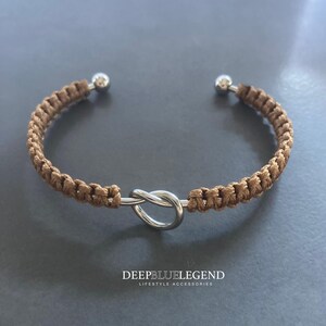 May include: A brown macrame bracelet with a silver knot design in the center. The bracelet has silver ball ends. The text "DEEPBLUELEGEND LIFESTYLE ACCESSORIES" is at the bottom.