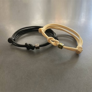 Può includere: Due braccialetti abbinati, uno nero e uno color crema. Ogni braccialetto ha una chiusura a nodo e una piccola perla a forma di cubo con una lettera. Il braccialetto nero ha la lettera "J", quello color crema ha la lettera "K".