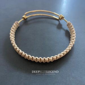 May include: A beige macrame bracelet with a gold-colored adjustable band. The macrame portion is tightly woven, and the bracelet is displayed on a gray surface. The text "DEEPBLUELEGEND LIFESTYLE ACCESSORIES" is visible.