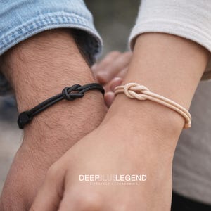 以下が含まれることがあります： 黒とベージュの、結び目デザインのペアブレスレット。 画像の下部には「DEEPBLUELEGEND LIFESTYLE ACCESSORIES」の文字が見えます。 手首に着用されています。