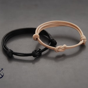 May include: Two adjustable bracelets. One is black, the other is beige. Each bracelet features a knotted design and is made of a thin, cord-like material. The bracelets are displayed on a gray surface.
