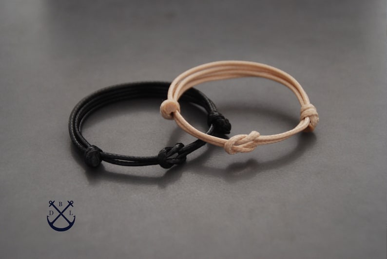 May include: Two adjustable bracelets. One is black, the other is beige. Each bracelet features a knotted design and is made of a thin, cord-like material. The bracelets are displayed on a gray surface.
