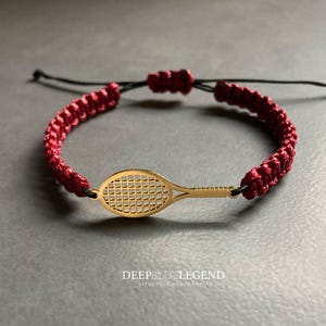 Puede incluir: Pulsera con temática de tenis con un dije de raqueta de tenis dorada. La pulsera tiene un cordón burdeos trenzado con lazos negros ajustables. El texto "DEEPBLUELEGEND LIFESTYLE ACCESSORIES" está en la parte inferior.