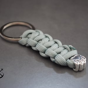Puede incluir: Un llavero de paracord verde claro con un dije de pata de plata. El llavero está hecho con un paracord trenzado y tiene un anillo de llave de plata.
