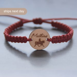 Op de afbeelding: Rood gevlochten armband met een houten bedel met de naam "Matthias" en een silhouet van een persoon die een paard rijdt.