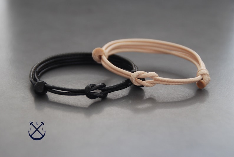 May include: Two adjustable bracelets. One is black, the other is tan. Each bracelet features a knotted closure and a simple, minimalist design. The bracelets are likely made of a durable, flexible material, suitable for everyday wear.