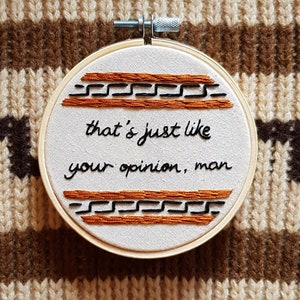 Könnte beinhalten: Stickrahmen-Kunst mit Stickerei und dem Text "that's just like your opinion, man" in schwarzer Kursivschrift auf weißem Hintergrund. Der Rahmen ist mit einem braunen und schwarzen Muster verziert.
