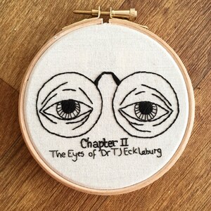 Puede incluir: Arte de bordado en aro con un par de ojos grandes con pupilas negras y pestañas, rodeado de un círculo blanco con el texto "Chapter II The Eyes of Dr. T.J. Eckleburg" en negro.