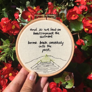 Puede incluir: Un bastidor de bordado redondo con la cita "and so we beat on boats against the current borne back ceaselessly into the past." El diseño incluye un muelle y una puesta de sol. El bastidor se sostiene contra un fondo de flores rojas.