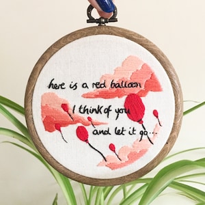 May include: Embroidered artwork in a wooden hoop. The design features red balloons and text that reads "here is a red balloon, I think of you and let it go..." The background is white fabric with a light pink cloud design.