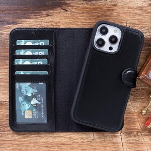 May include: Black leather phone case with a built-in wallet. The case has slots for credit cards and a flap that covers the phone screen. The phone case is lying on a wooden surface.