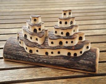 May include: A handcrafted wooden toy boat with multiple levels and small, dark-colored openings. The boat is made from light-colored wood with darker, burnt-looking edges, and sits on a wooden surface.