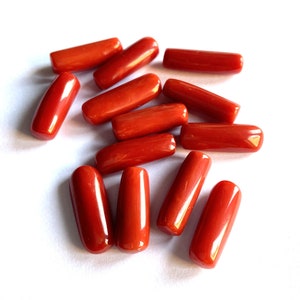 Puede incluir: Una colecci&oacute;n de gemas de coral rojo pulido y vibrante. Cada gema tiene una forma ovalada alargada y lisa. Las piezas de coral est&aacute;n dispersas sobre una superficie blanca, mostrando su rico color y belleza natural.