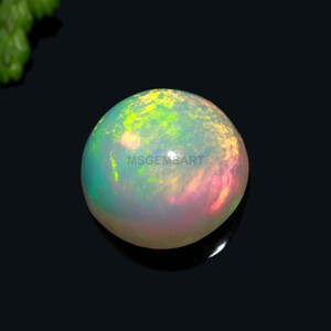 Puede incluir: Una piedra preciosa de ópalo redonda y blanca con destellos verdes y rosados. El ópalo tiene un diámetro de aproximadamente 10 mm.