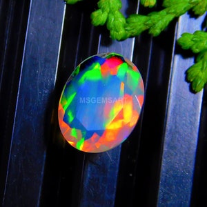 Puede incluir: Una gema ovalada con una vibrante exhibición de colores, incluyendo verde, azul, rojo y amarillo. La gema está facetada, creando múltiples superficies reflectantes. El texto "MSGEMSART" es visible en la gema. El fondo es oscuro con algunas hojas verdes.