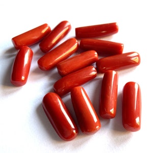 Puede incluir: Una colecci&oacute;n de cuentas de coral rojo pulido y vibrante, cada una con una forma cil&iacute;ndrica y lisa. Las cuentas est&aacute;n dispersas sobre una superficie blanca, mostrando su rico color y brillo natural. Ideales para la creaci&oacute;n de joyas.