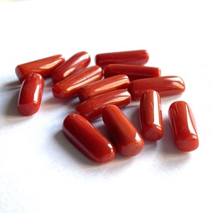 Puede incluir: Una colecci&oacute;n de piedras preciosas de coral rojo pulido, de forma ovalada. Las piedras son de un rojo vibrante y brillante, dispersas sobre una superficie blanca. Las piezas de coral var&iacute;an ligeramente en tama&ntilde;o y forma, con extremos redondeados.