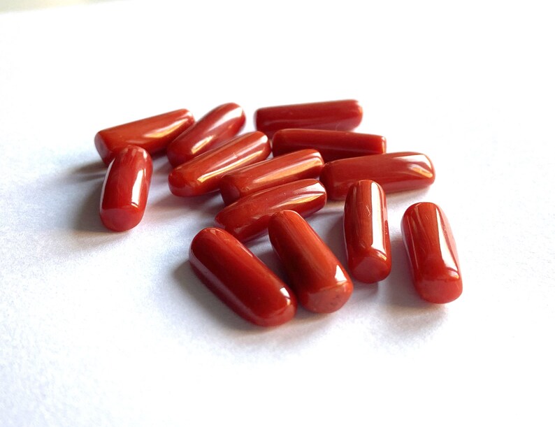 Puede incluir: Una colecci&oacute;n de piedras preciosas de coral rojo pulido, de forma ovalada. Las piedras son de un rojo vibrante y brillante, dispersas sobre una superficie blanca. Las piezas de coral var&iacute;an ligeramente en tama&ntilde;o y forma, con extremos redondeados.