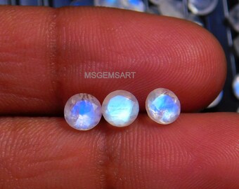 May include: Three round, iridescent moonstone gemstones with a blue sheen. The stones are approximately 6mm in diameter and are arranged in a row on a finger.