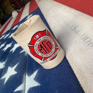 May include: A white can cooler with a red circle logo that says "Milwaukee Fire Department - Established 1875 - Courage - Integrity - Honor". The logo has a red star and a red cross inside the circle.