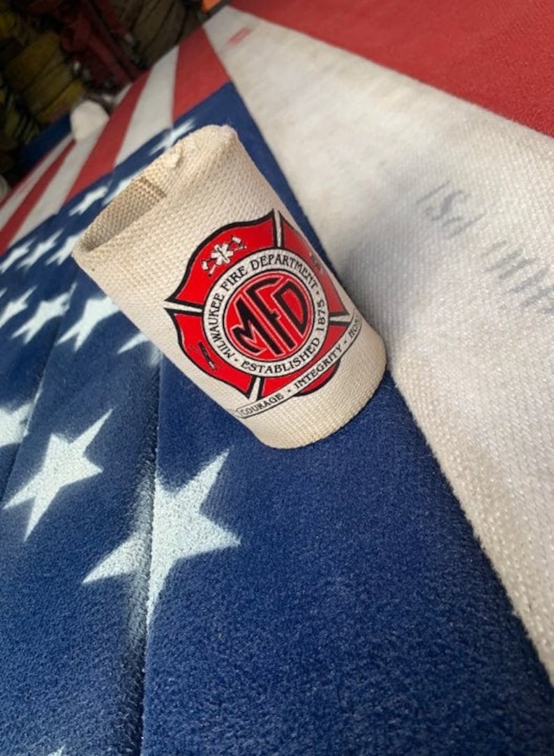 May include: A white can cooler with a red circle logo that says "Milwaukee Fire Department - Established 1875 - Courage - Integrity - Honor". The logo has a red star and a red cross inside the circle.