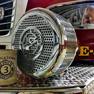 May include: A close-up of a chrome fire truck siren with a diamond plate surface in the background. The siren has a large, circular grill with a smaller, circular logo in the center. The fire truck has a yellow "E-31" designation on the side.