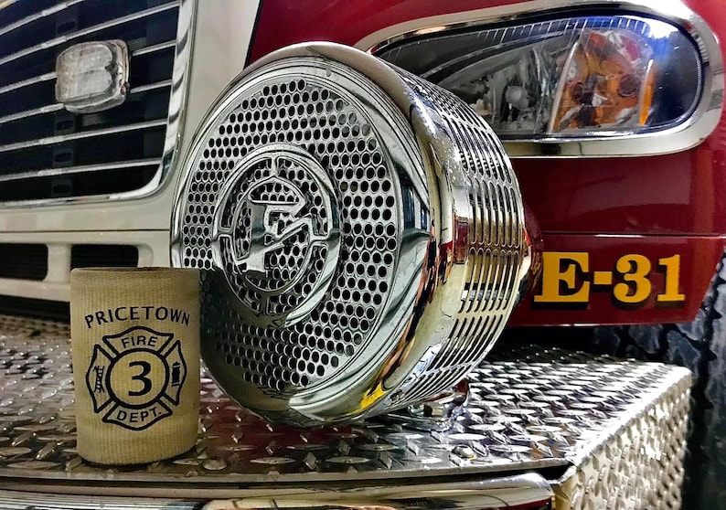 May include: A close-up of a chrome fire truck siren with a diamond plate surface in the background. The siren has a large, circular grill with a smaller, circular logo in the center. The fire truck has a yellow "E-31" designation on the side.