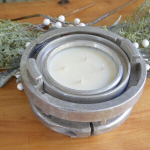 May include: A white candle with four wicks sits inside a silver metal fire hydrant-shaped candle holder. The candle holder is on a wooden surface with green foliage and white berries.