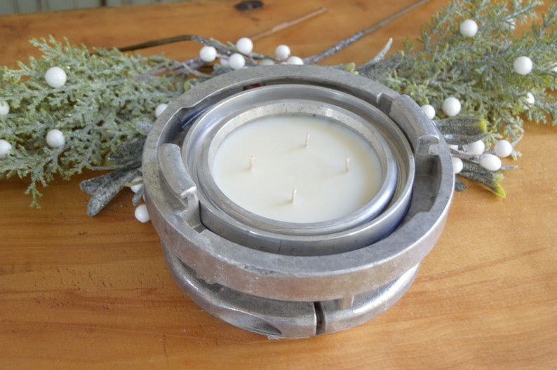 May include: A white candle with four wicks sits inside a silver metal fire hydrant-shaped candle holder. The candle holder is on a wooden surface with green foliage and white berries.