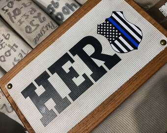 May include: A wooden sign with the word "HER" in large black letters. A blue line American flag patch is in the upper right corner of the sign.
